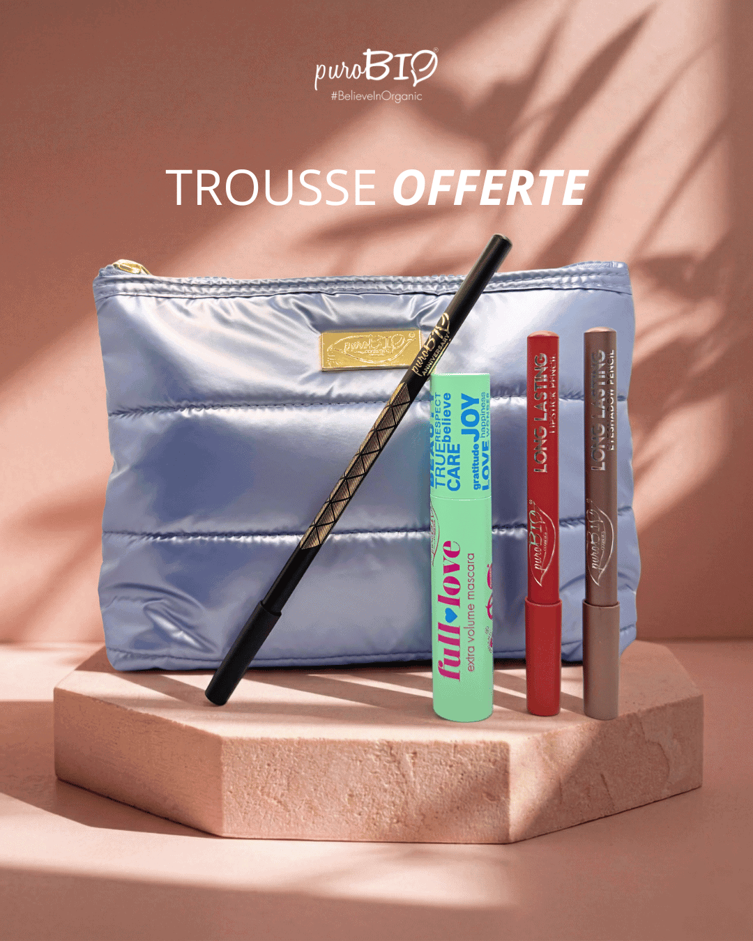 Trousse de maquillage argentée puroBIO offerte contenant un mascara vert, un crayon noir pour les yeux et deux crayons longue tenue pour les lèvres et le teint, posée sur un socle beige avec fond rose.
